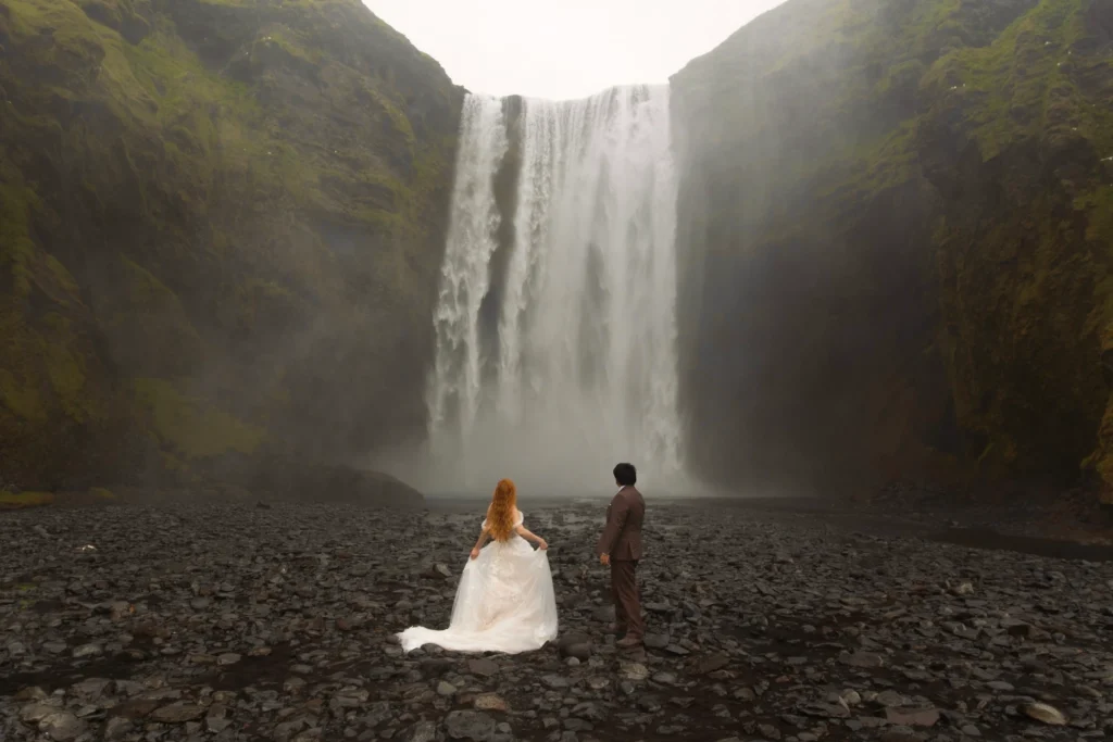A bride and Groom stand near the base of a waterfall.