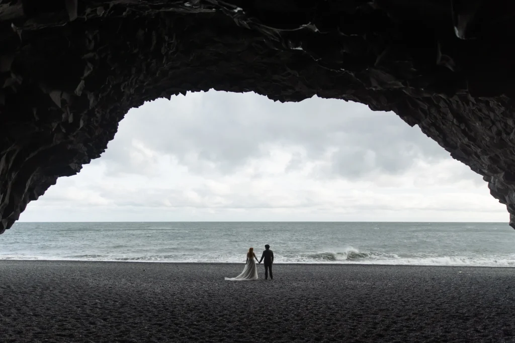 a bride and groom stand on the beach, framed by a large cave