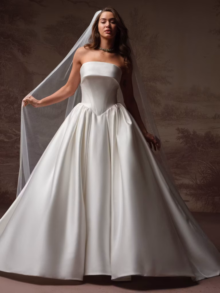 Rane by Maggie Sottero satin ball gown wedding dress featuring a basque waist, strapless straight neckline, hidden pockets, and a dramatic full skirt.